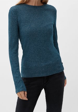 Kvinde iført en tætsiddende, langærmet, blågrøn ribstrikket trøje sammen med sorte bukser, stående foran en ensartet lysegrå baggrund.