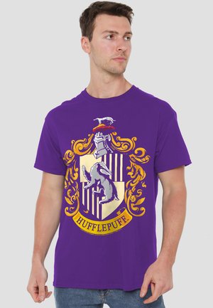 Young man wearing a purple T-shirt with a large yellow and black Hufflepuff crest from Harry Potter, looking to his right.