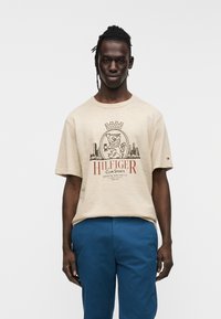 Μπεζ βαμβακερό t-shirt με τυπωμένο σχέδιο με κονκάρδα και λιοντάρι, κείμενο «HILFIGER CLUB SPORTS» κόκκινο. Το μοντέλο φοράει μπλε παντελόνι.