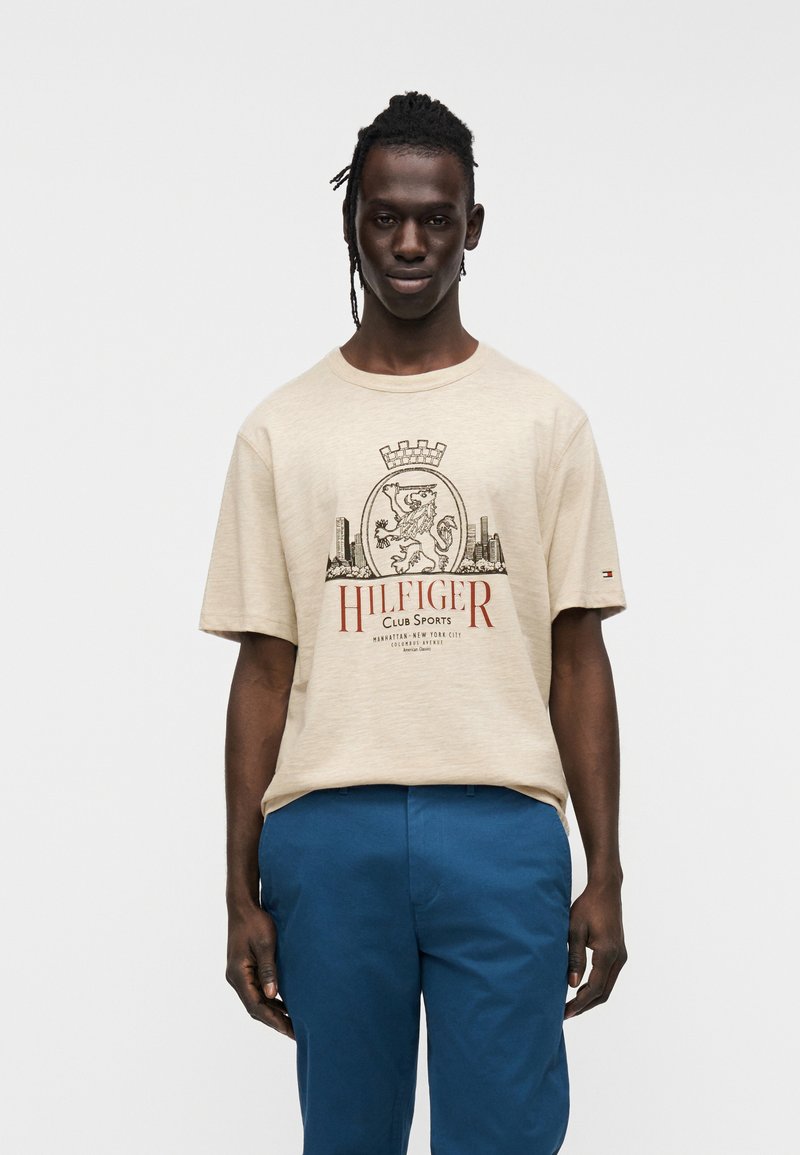 Μπεζ βαμβακερό t-shirt με τυπωμένο σχέδιο με κονκάρδα και λιοντάρι, κείμενο «HILFIGER CLUB SPORTS» κόκκινο. Το μοντέλο φοράει μπλε παντελόνι.