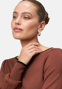 Brązowy sweter z czarnymi wykończeniami, z bufiastymi rękawami. Modelka nosi srebrne kolczyki w kształcie kół i ma starannie związane włosy w kok.