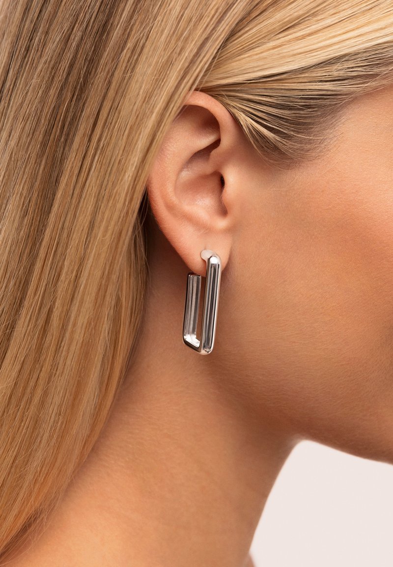 Pendientes geométricos plateados con un acabado brillante. Presentan formas rectangulares alargadas con un diseño elegante. Llevados en una oreja.