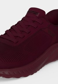 Σκούρο κόκκινο slip-on sneaker Skechers με πλεκτό πάνω μέρος, ελαστικά κορδόνια και υφασμένες πλευρές, εμφανίζεται σε λευκό φόντο.