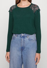 Dark green long-sleeve top featuring lace panels on the shoulders, paired with high-waisted light blue denim jeans.