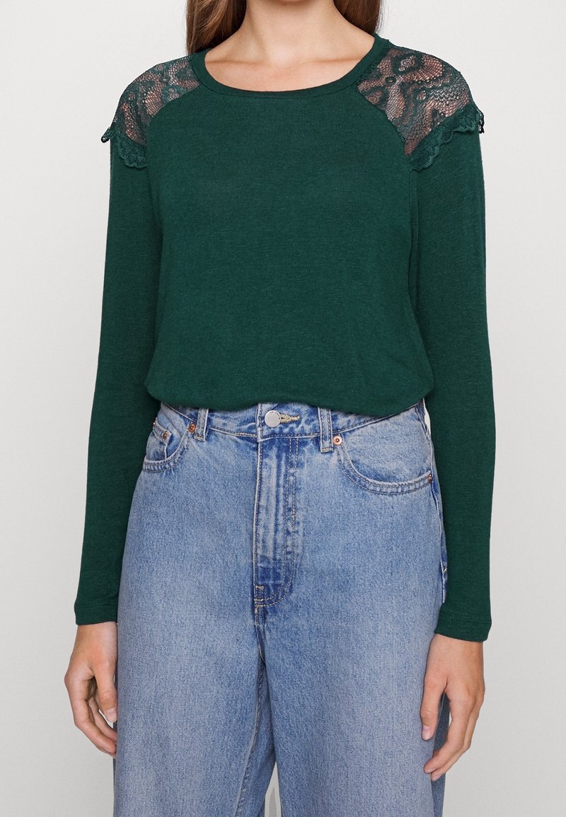 Dark green long-sleeve top featuring lace panels on the shoulders, paired with high-waisted light blue denim jeans.