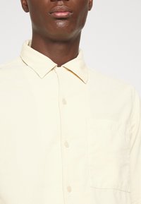 Jasnobeżowa koszula na guziki z klasycznym kołnierzykiem, kieszonką na piersi i subtelną fakturą tkaniny. Charakteryzuje się klasycznymi guzikami i czystymi liniami.