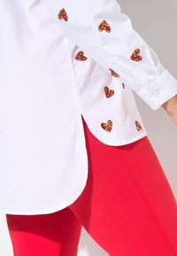 Camicia bianca con applicazioni a forma di cuore rosso lucido sulle maniche, abbinata a leggings rossi aderenti. Caratterizzata da un orlo arrotondato e polsini con bottoni.