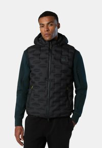 Chaleco acolchado negro con textura de rombos, capucha ajustable, cierre frontal con cremallera y bolsillos laterales. Llevado sobre una camiseta de manga larga verde oscuro.