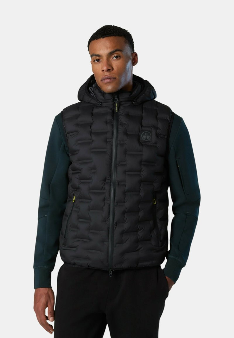 Chaleco acolchado negro con textura de rombos, capucha ajustable, cierre frontal con cremallera y bolsillos laterales. Llevado sobre una camiseta de manga larga verde oscuro.