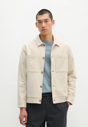 Man draagt een beige jas met knopen en grote borstzakken over een blauw hemd en een donkere broek, staand tegen een effen witte achtergrond.