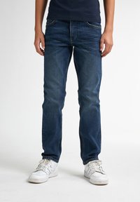 Jean en denim bleu à coupe droite, avec cinq poches et des détails de whiskering subtils. Porté avec des sneakers de sport blanches.