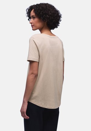 Donna con capelli ricci che indossa una maglietta beige a maniche corte e pantaloni neri, vista di profilo con uno sfondo neutro.