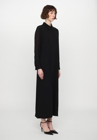 Vestido negro de manga larga con cuello, diseño de pliegues en el frente, textura suave, hasta los tobillos, combinado con tacones de aguja negros y correas en los tobillos.