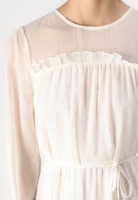 White dress with a textured, sheer overlay, ruffled neckline, and metallic accents. Features long sleeves and a cinched waist.