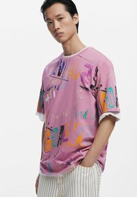 Roze oversized T-shirt met meerkleurige abstracte graphics, ronde halslijn en korte witte mouwen. Gecombineerd met gestreepte broek.