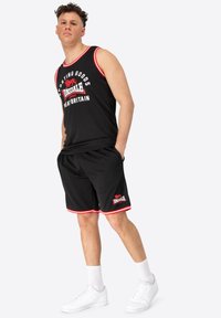 Čierny športový tank top s červenými akcentmi a logom, v kombinácii s čiernymi šortkami s červeným lemovaním. Nosí sa so systémami bielymi teniskami.