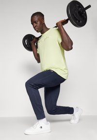 Lichtgroene t-shirt met 'Champion' logo, marineblauwe joggingbroek, witte sneakers en een zwarte halterstang op de schouders in een workout houding.