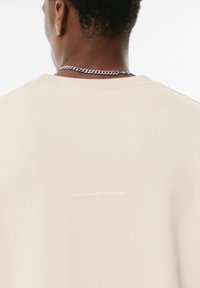 Beige sweatshirt med rund hals, med subtil tekst på ryggen, der står: "Jeg tror ikke, dette er min plads." Blød stofstruktur.