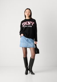 Černý pletený svetr s nápisem "DKNY NEW YORK" v červené a bílé barvě, sladěný se světle modrou džínsovou mini sukní a černými vysokými botami po kolena.