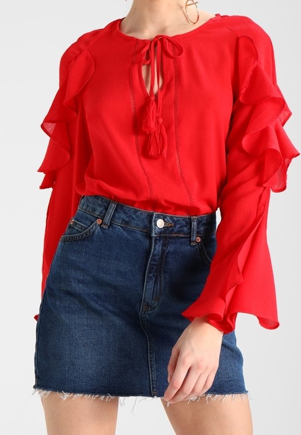 Femme portant un chemisier rouge vif avec des manches volantées et des liens à pompons, associé à une mini-jupe en jean bleu foncé avec un ourlet effiloché.
