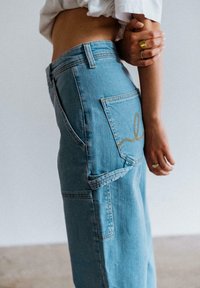 Hellblaue Jeans mit entspannter Passform, versehen mit einem goldenen Stickdetail an der Gesäßtasche und einer sichtbaren Seitennaht.