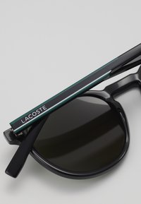 Juodos saulės akinių su blizgiu paviršiumi, turinčių ryškų Lacoste logotipą žaliame ir juodame dryžuotame išsikišančiame krašte, ir tamsiais lęšiais.