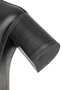 Zapato de tacón alto negro con parte superior de cuero suave y un tacón grueso y texturizado que presenta una base redondeada y un acabado mate.