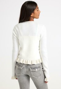 White, fluffy sweater with ribbed hem and cuffs, featuring a ruffled edge. Paired with light gray jeans having detailed back pockets.