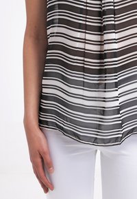 Gros plan d'une personne portant un haut transparent rayé noir et blanc avec un pantalon blanc, montrant le bras gauche et la main détendus sur le côté.