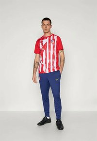 Uomo che indossa una maglietta da calcio dell'Atlético Madrid a strisce rosse e bianche, pantaloni da corsa Nike blu e scarpini neri, in piedi davanti a uno sfondo semplice.