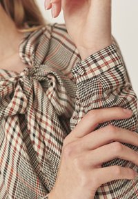 Nööbiga särk, millel on ruuduline muster beeži, musta ja roostespruuni värviga, kinni seotud kaelaosa ja struktureeritud manseti detailid.