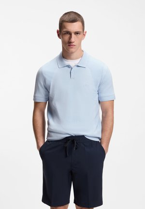 Junger Mann steht mit den Händen in den Taschen, trägt ein hellblaues Poloshirt mit kurzen Ärmeln und marineblaue Shorts mit Kordelzug vor weißem Hintergrund.