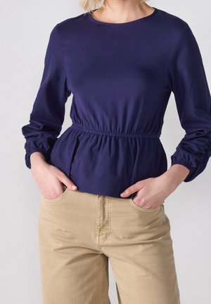 Kobieta ubrana w granatową bluzkę z długim rękawem i elastycznym pasem, w połączeniu z beżowymi spodniami, z rękami włożonymi do kieszeni.