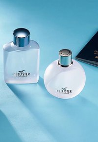 To flasker med Hollister-duft på en blå bakgrunn. Venstre: rektangulært frostet glass med en blå metallisk hette. Høyre: rundt frostet glass med en sølvhette.