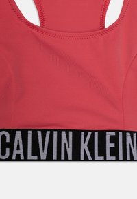Grimizni sportski grudnjak s glatkom teksturom, s širokim crnim pojasom na kojem stoji "CALVIN KLEIN" u podebljanim bijelim slovima.