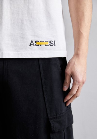 Hvit bomull T-skjorte med svart og gul tekstlogo "ASPRISI" nederst. Modellens hånd hviler på sorte cargo shorts.
