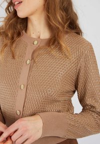 Jasnobrązowy sweter wykonany na drutach z teksturowanym wzorem w diamenty, złotymi guzikami i prążkowanymi mankietami. Zbliżenie na górną część.