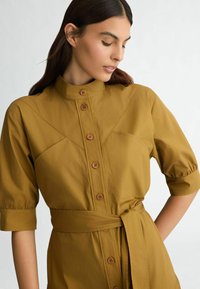 Robe longue en moutarde avec un col haut, un devant à boutons et de courtes manches bouffantes. Caractérisée par une ceinture à la taille et des poches au niveau de la poitrine.