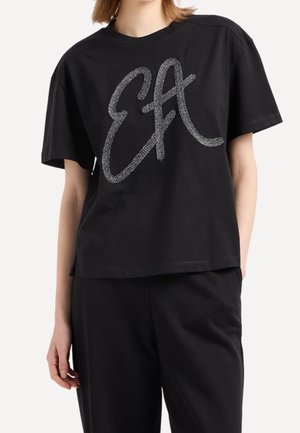 Personne portant un t-shirt noir à manches courtes avec de grandes lettres argentées pailletées "EA" à l'avant et un pantalon noir.