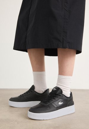 Sneakers in pelle nera con suola in gomma bianca, dotati di un logo di lato, punta rotonda e lacci neri. Indossati con calze bianche.