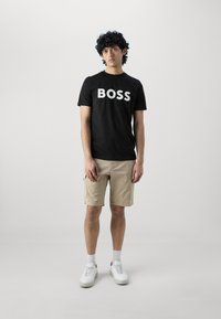 Melnas kokvilnas t-krekls ar lielu baltu "BOSS" logo, bēšas cargo šortus, baltas kedas un pelēkas zeķes, stāvot pret vienkāršu fona.