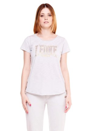 Une femme porte un T-shirt clair avec l’inscription dorée « LEONE » et un pantalon assorti, sur fond blanc.