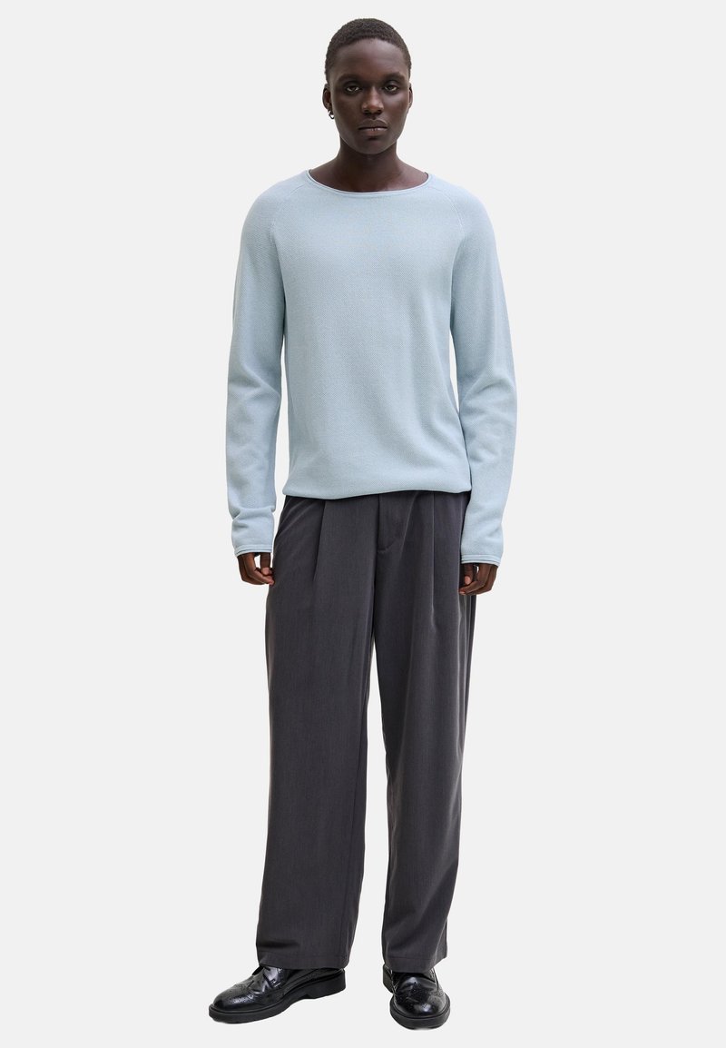 Personne debout portant un pull à manches longues bleu clair, un pantalon ample gris foncé et des chaussures habillées noires sur un fond blanc.
