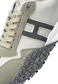 Zapatilla perforada gris claro con acentos de gamuza verde apagado, que presenta cordones beige, una suela de goma y un parche con el logo en el costado.