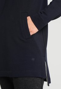 Túnica de manga larga en color navy, hecha de tela suave, con un bolsillo lateral, aberturas en los laterales y un pequeño logo en el dobladillo. Textura suave.