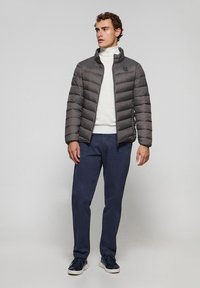 Grå puffjacka med horisontell quiltníng, hög krage och logopatch, kombinerad med en vit polo, marinblå byxor och mörka sneakers.