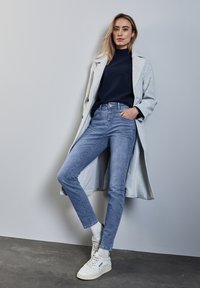 Ljusgrå lång kappa över en marinblå polotröja, tillsammans med ljusblå jeans och vita högtoppsneakers på en mörk yta.