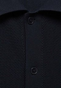 Námořnicky modré polo tričko s texturovanou tkaninou, malým límcem a dvěma černými knoflíky vpředu.