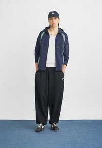 Fleece com fecho éclair azul-marinho com detalhes em verde, combinado com calças de treino pretas largas e ténis pretos. T-shirt branca por baixo e boné preto utilizado.