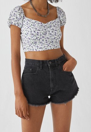 Femme portant un top court blanc à fleurs avec des manches bouffantes et un short en jean noir effiloché, une main dans la poche.